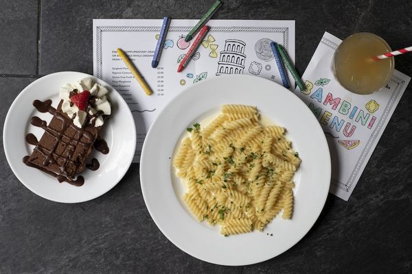 A plate of pasta and a portion of chocolate cake with sauce and cream. Served alongside kids colouring pages and crayons.