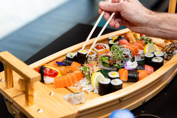 hand with chopsticks picking up sushi from a small wooden boat serving platter.