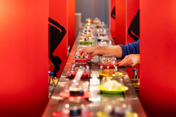 a silver conveyer belt with little sushi dishes flanked by red panels. a hand taakes two dishes from the conveyer belt