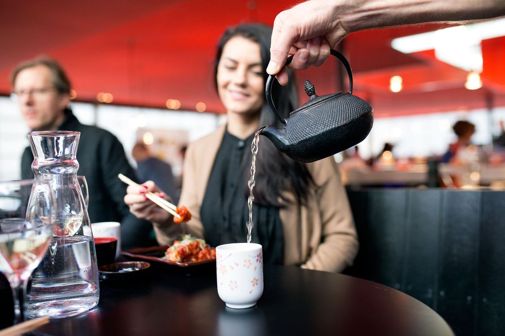 Serving-Green-Tea-at-Moshimo-Restaurants-Brighton. Pictured an attractive young lady sat at a Japanese style table. the table is plack, there is a water jug in the foreground and a blurred table guest in the background. You can see the red ceiling canape. 