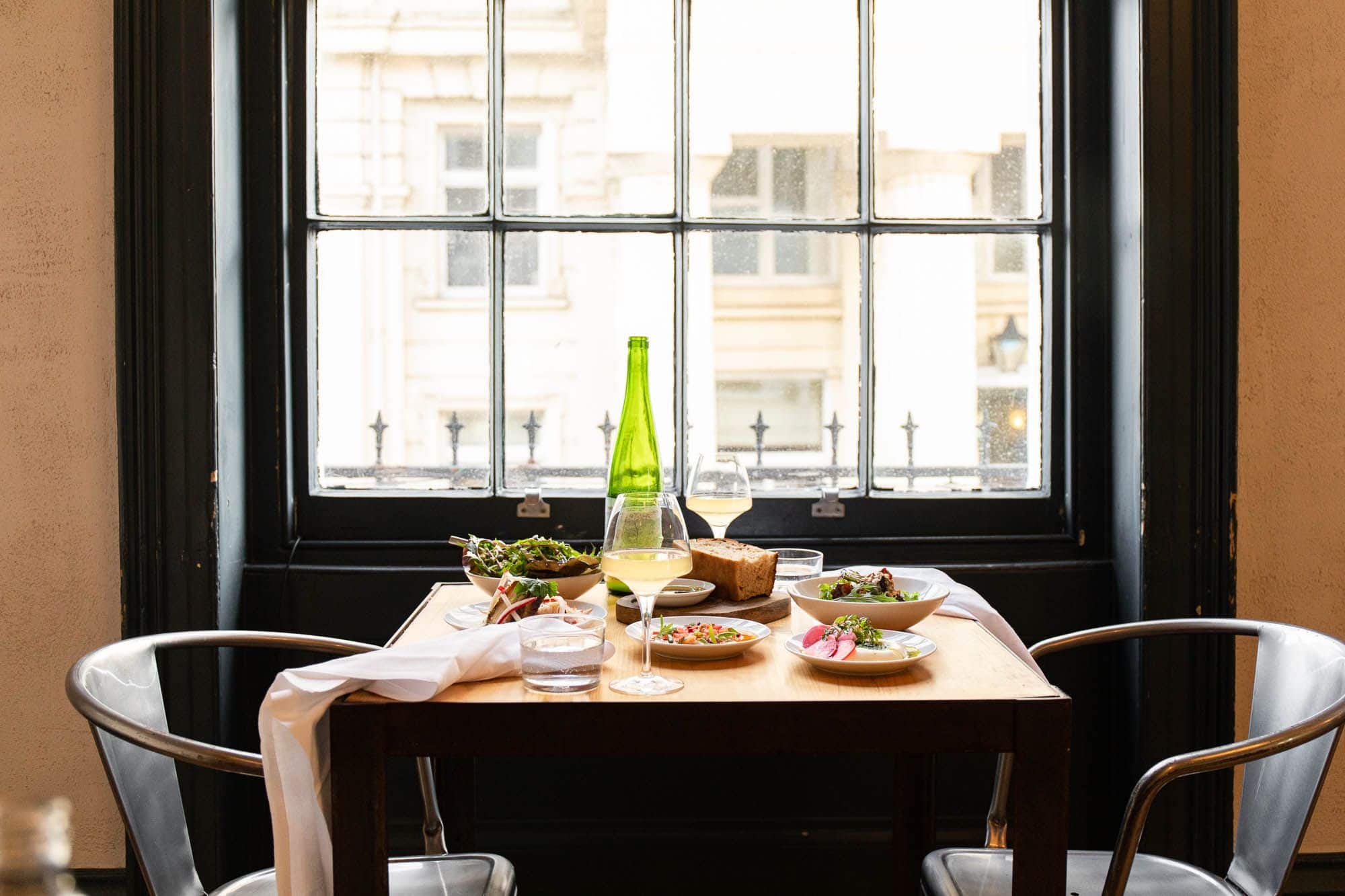 A table for two with plates of food and a bottle of wine with two wine glasses full of white wine. Shot in front of the window with a view of the street.