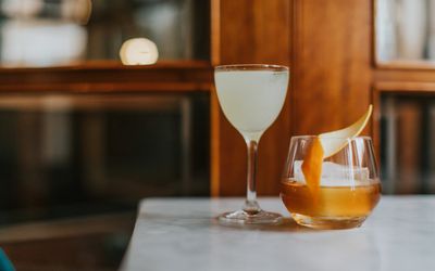 Two cocktails on the edge of a table with a wooden backdrop. One short glass with a fruit crisp garnish and one taller glass with a different drink.