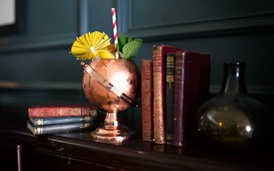 A copper globe shaped cocktail glass with a red and white paper straw and paper cocktail umbrella decoration. Sat on a book case.