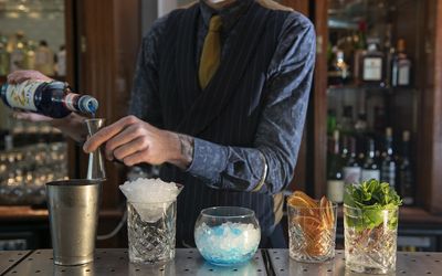 Bartender preparing cocktails with different glasses and garnishes. Bars in Brighton. There are four cocktails on display and a cocktail mixer. The bartender is filling his measuring cup.