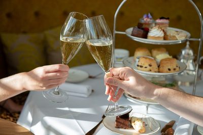 Two hands holding champagne flutes with a three tier cake stand in the background full of cakes and sandwiches.