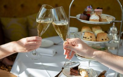 Two hands holding champagne flutes with a three tier cake stand in the background full of cakes and sandwiches.