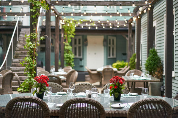 Sage green outdoor area with woonden pagoda, fairy lights, wicker seating and tables dressed with glasses and red flowers.