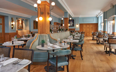 a large duck egg blue dining rooms with several tables and chairs with banquetter seating in the centre. The floor is wooden and the tabletops are marble.