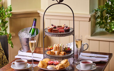 a three tiered caake plate with cakes on the top layer, sandwiches and nibbles on the middle and scones with jam and cream on the bottom. Table has champagne and cups of tea.