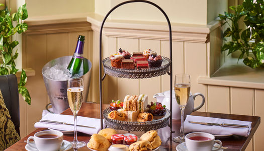 a three tiered caake plate with cakes on the top layer, sandwiches and nibbles on the middle and scones with jam and cream on the bottom. Table has champagne and cups of tea. Afyernoon Tea in Brighton