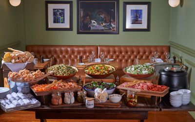 A large table with buffet food on top. Bowls of salad. Platters of meat, cheeses and fish. The table sits infront of leather bench and green wall.