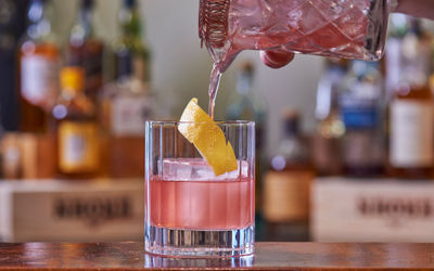 pink cocktail drink being poured into a short cut glass. Lemon peel twist in the glass. Bottles in the background