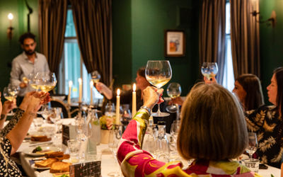 In a dark green room is a table filled with people who are tasting wine. There is a guide who is asking all the diners to lift up their glasses to look at something wine related.