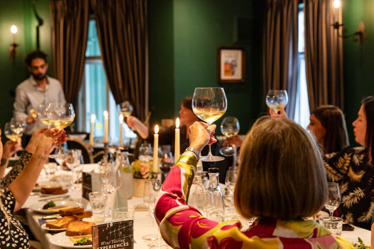 In a dark green room is a table filled with people who are tasting wine. There is a guide who is asking all the diners to lift up their glasses to look at something wine related.