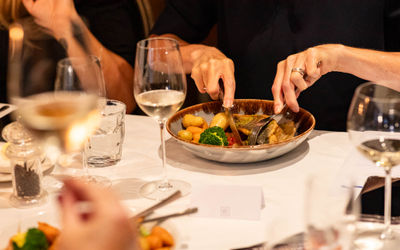 The setting is a dining table with a white cloth and glasses. We see a close up of hands using a fork and knife as they dine on a fish dish. It seems formal yet relaxed.