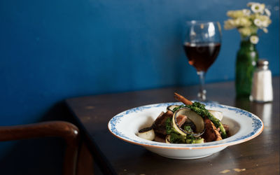 A main dish served on the table with a glass of red wine and a vase of flowers. Photographed against a dark blue wall.