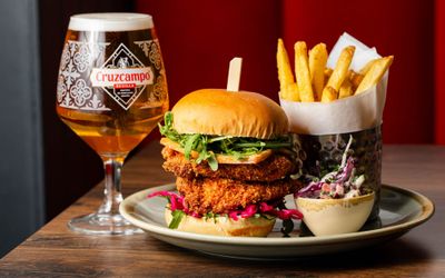 Crispy fried chicken burger stacked with salad, served with golden fries, slaw, and a glass of Cruzcampo lager.