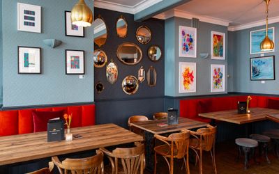 Cozy restaurant interior with wooden tables, red cushioned seating, decorative mirrors, and colorful framed artwork on the walls.