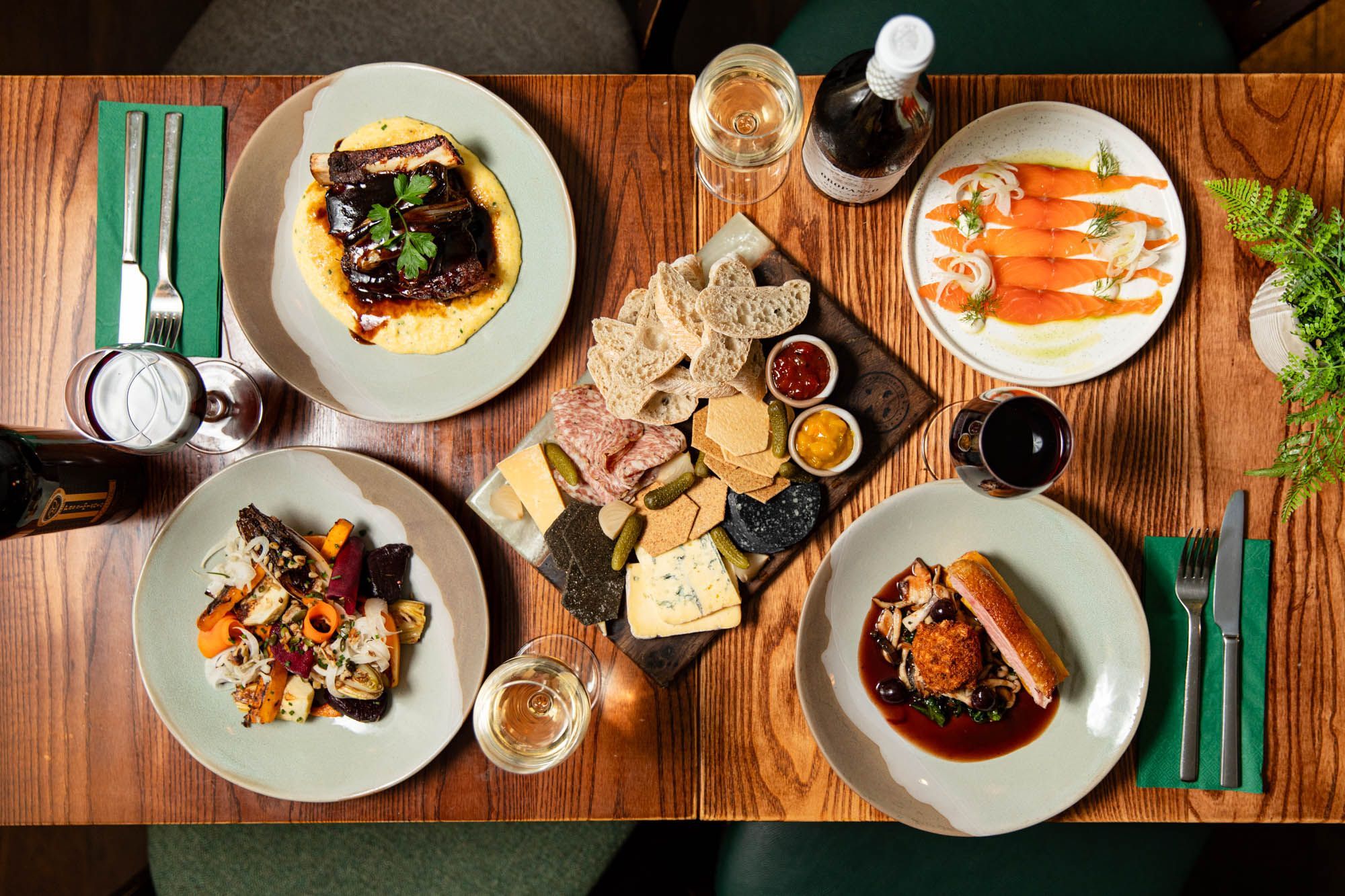 over head shot of the wooden brown table laid out with four different plates of food and cheese and charcuterie board in the middle