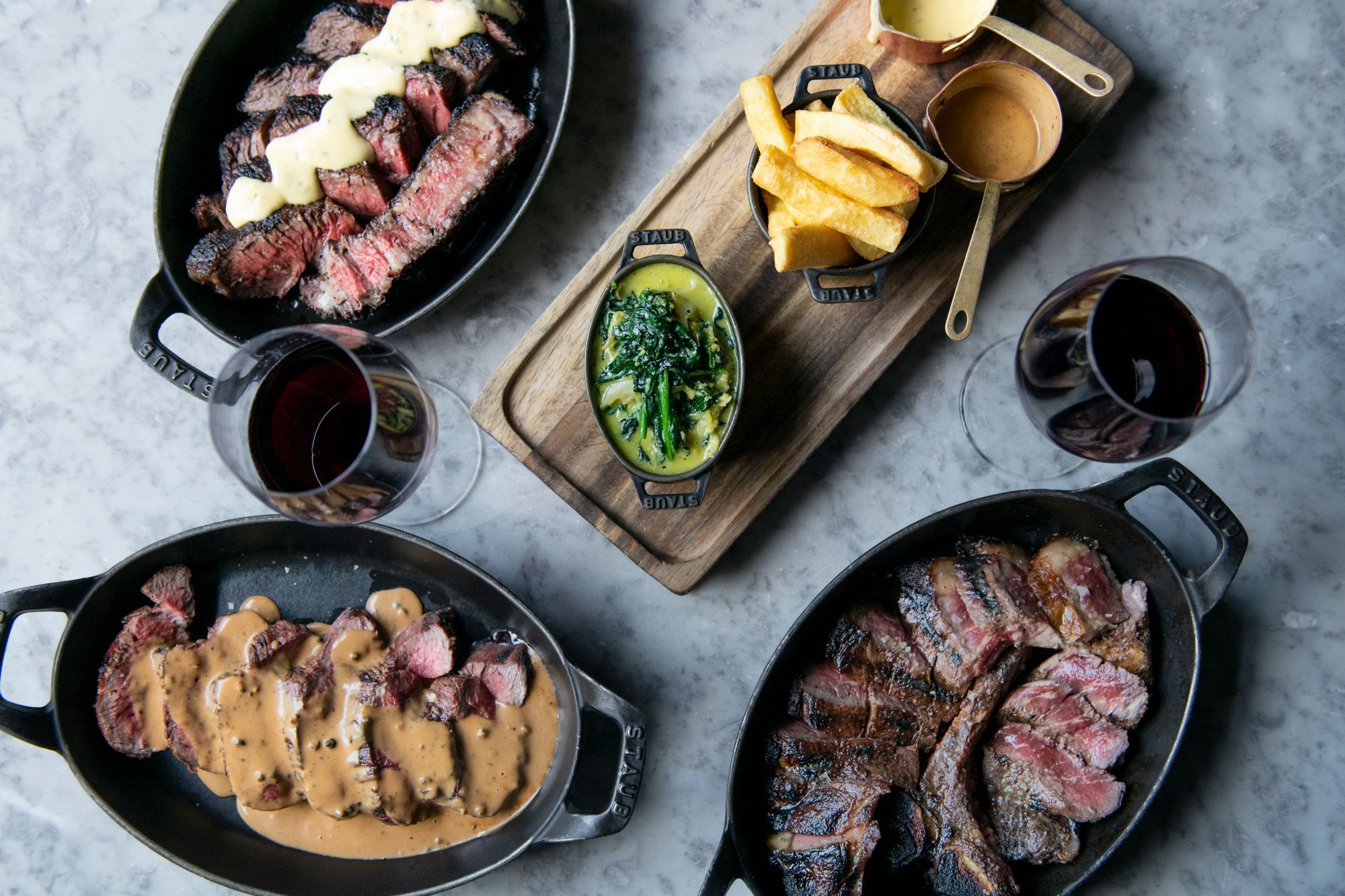 An overhead shot of a table of food. Three black iron dishes with sliced steak cooked rare and seared to perfection. Two glasses of red wine and a board with chips and vegetables accompany the meal.