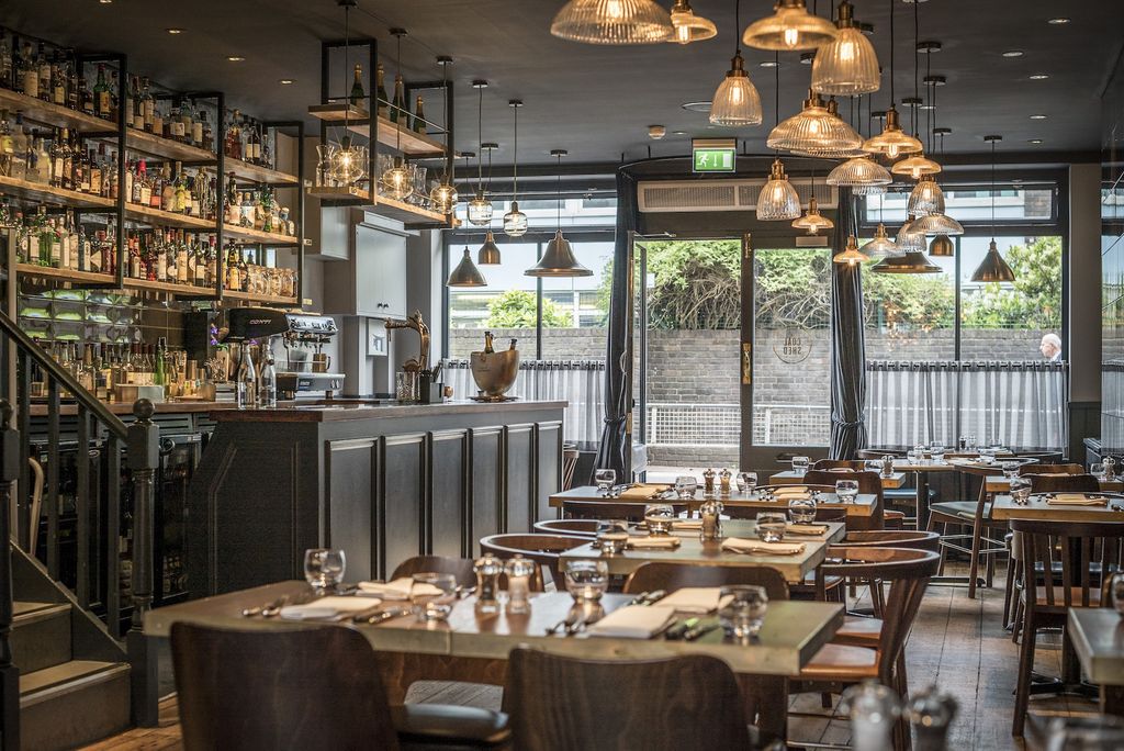 Bright and airy daytime shot of The Coal Shed Brighton interior. Showing tables set out for service, a number of glass ceiling lamps of various shapes and sizes and a well-stocked bar.