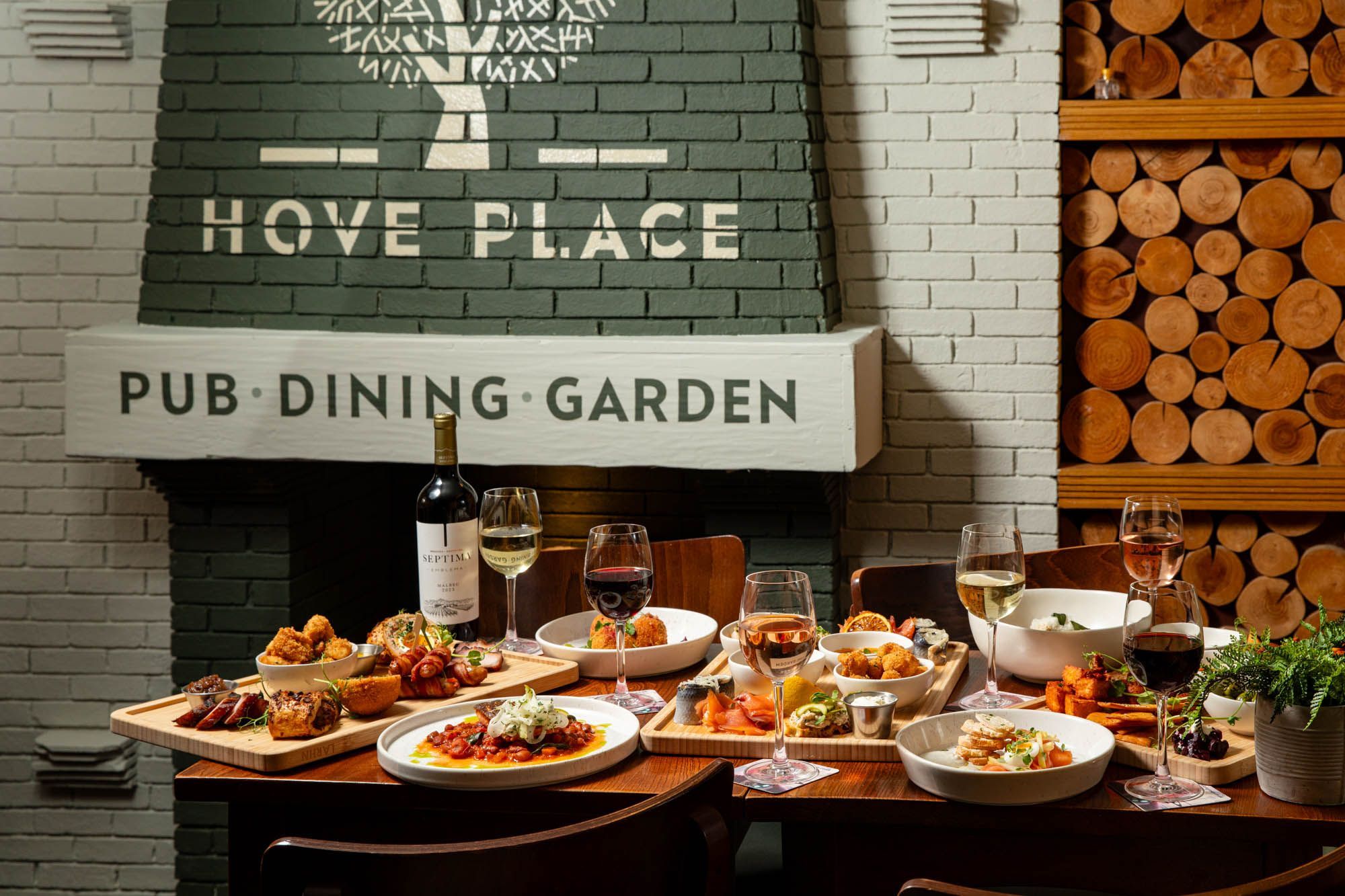 A beautifully set dining table at Hove Place, featuring an array of dishes including charcuterie boards, seafood, and sharing plates, accompanied by glasses of red, white, and rosé wine. The cozy pub setting includes a fireplace with the "Hove Place Pub Dining Garden" sign, a wood log feature wall, and warm lighting, creating an inviting and stylish dining atmosphere.