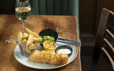 Fish and chips served on a blue ceramic plate with a pot of mushy peas, tartare sauce and a grilled lemon half. Served with a glass of white one on a wooden table.
