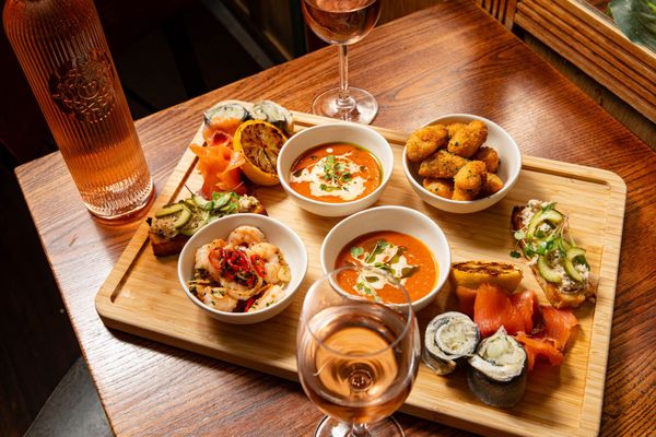 This image showcases a beautifully arranged seafood and tapas platter served on a wooden board. The spread includes bowls of rich tomato-based soup garnished with fresh herbs and cream, golden crispy bites, smoked salmon, marinated prawns with red chili, grilled lemon, and small bites of seafood-topped bruschetta. Accompanied by a bottle of elegant rosé wine and glasses of wine, this dish highlights a luxurious dining experience, perfect for sharing. The warm wooden table setting and soft lighting create an inviting and sophisticated atmosphere.