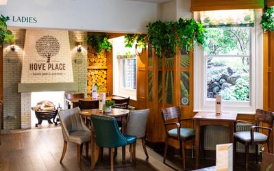 Interior of the hove place pub which includes fireplace, couple of brown tables and different colored chairs, plants on the wall and cosy lights