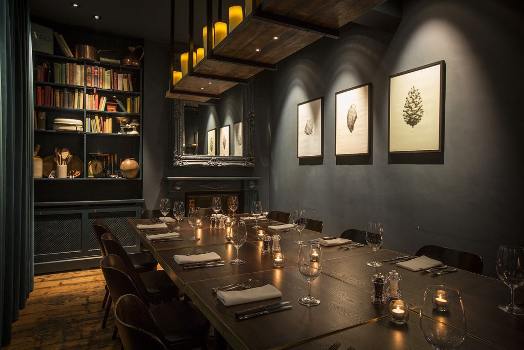 Dark grey painted room with a bookcase on one wall and three black and white drawings on the other. A long rectangular table is laid out for private dining. There are candles on the table. The space looks warm, sophisticated and elegant.