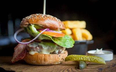 Steak burger stacked with red onion & tomato slices, bacon and cheese. Served alongside a gherkin and chunky chips on a wooden board.