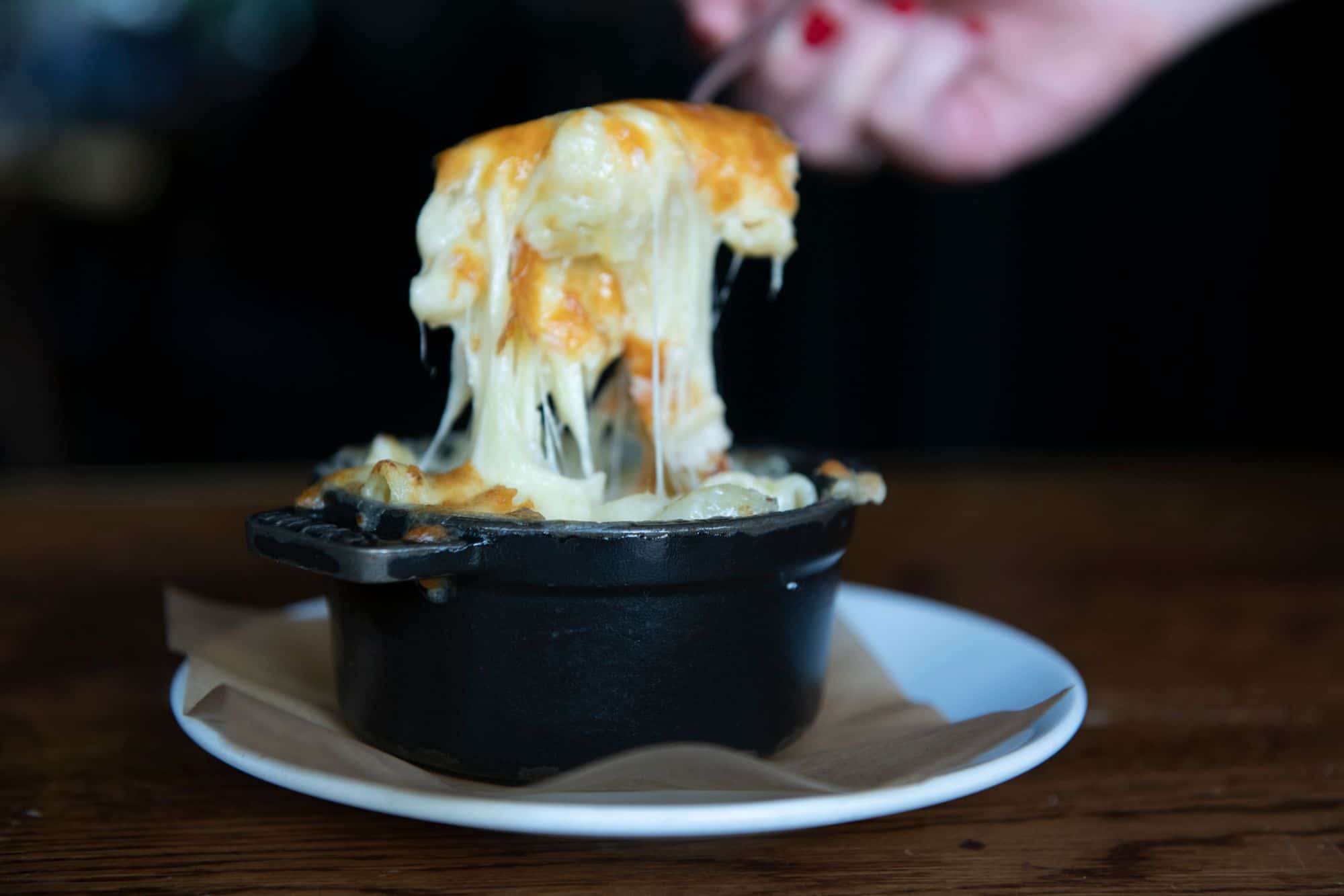The melt in the mouth short-Rib Mac n’ Cheese at the Coal Shed Brighton. A hand with red painted nails holds up a fork full of food with molten cheese dripping stretching from the black iron dish below.