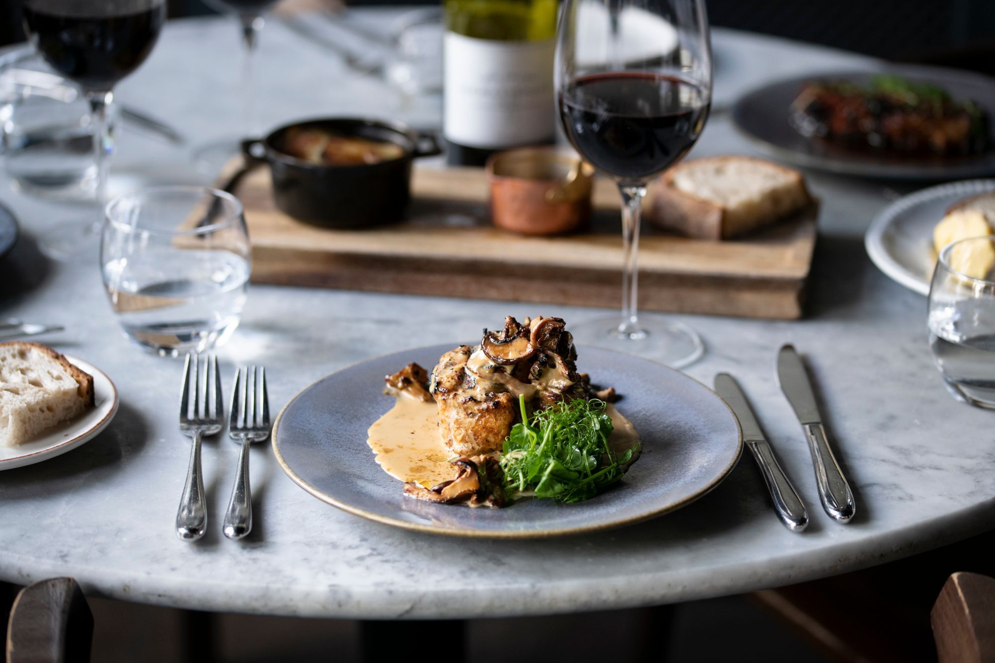 A monkfish dish, plated up with some crisp fresh greens and an orange coloured, creamy sauce. We see a glass of red wine and other dishes are blurred in the background.