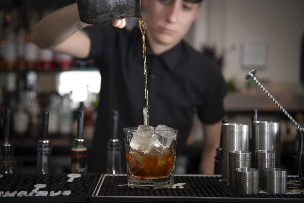 A mixologist in a blak shirt is at a granite bar mixing a cocktail