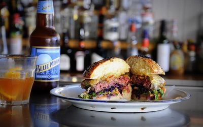 A photo of a beef burger cut in half on a bar with a bottle of Blue Moon beer and drinks in the background.