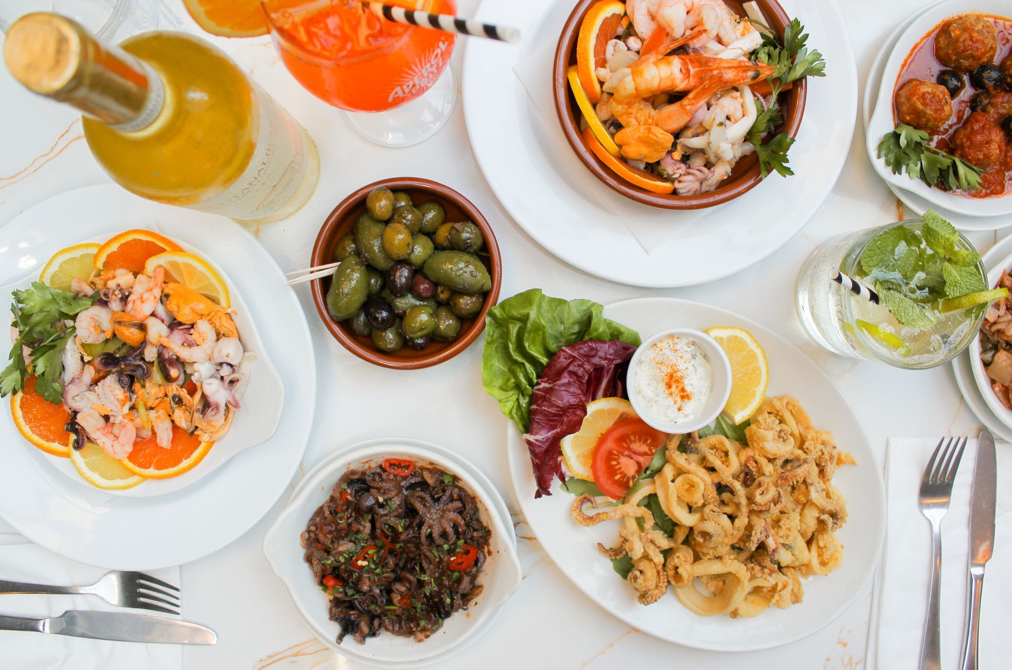 An over head shot of a white marble table laid out with plates of food, a seafood salad on a bed of citrus fruit, a side bowl of olives, peppered battered squid with salad, king prawns and meat balls. There are cocktails and a bottle of wine on the talble.