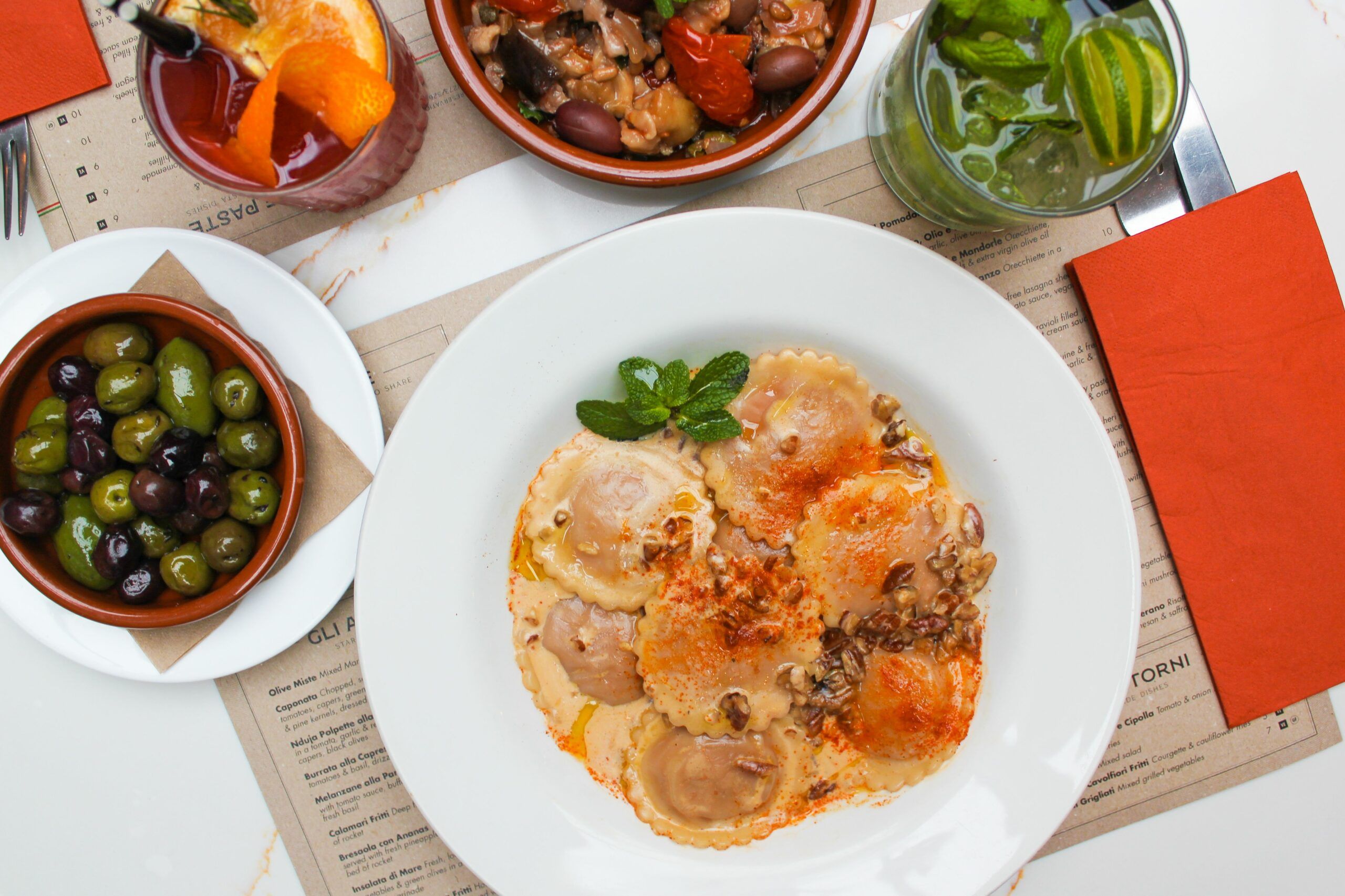 over head shot of raviolli in a round white pasta bowl at Al duomo Brighton. Around the ravioli there are bowls of olives, a side dish with beans a green drink with lime and mind and a red drink with citrus fruit garnish