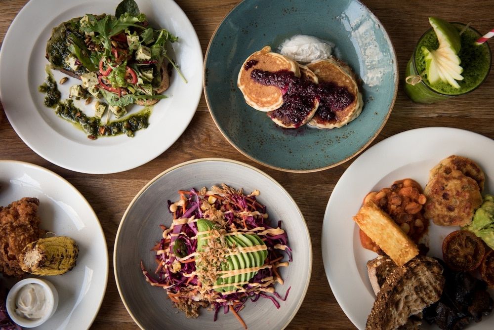 A selection of brunch meals laid out on a wooden table. Including an avocado salad and blueberry pancakes.
