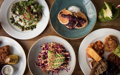 A selection of brunch meals laid out on a wooden table. Including an avocado salad and blueberry pancakes.