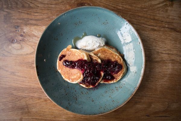 American style pancakes with a blueberry compote and cream, drizzled with maple syrup. Served in a duck egg blue ceramic dish on a wooden table.