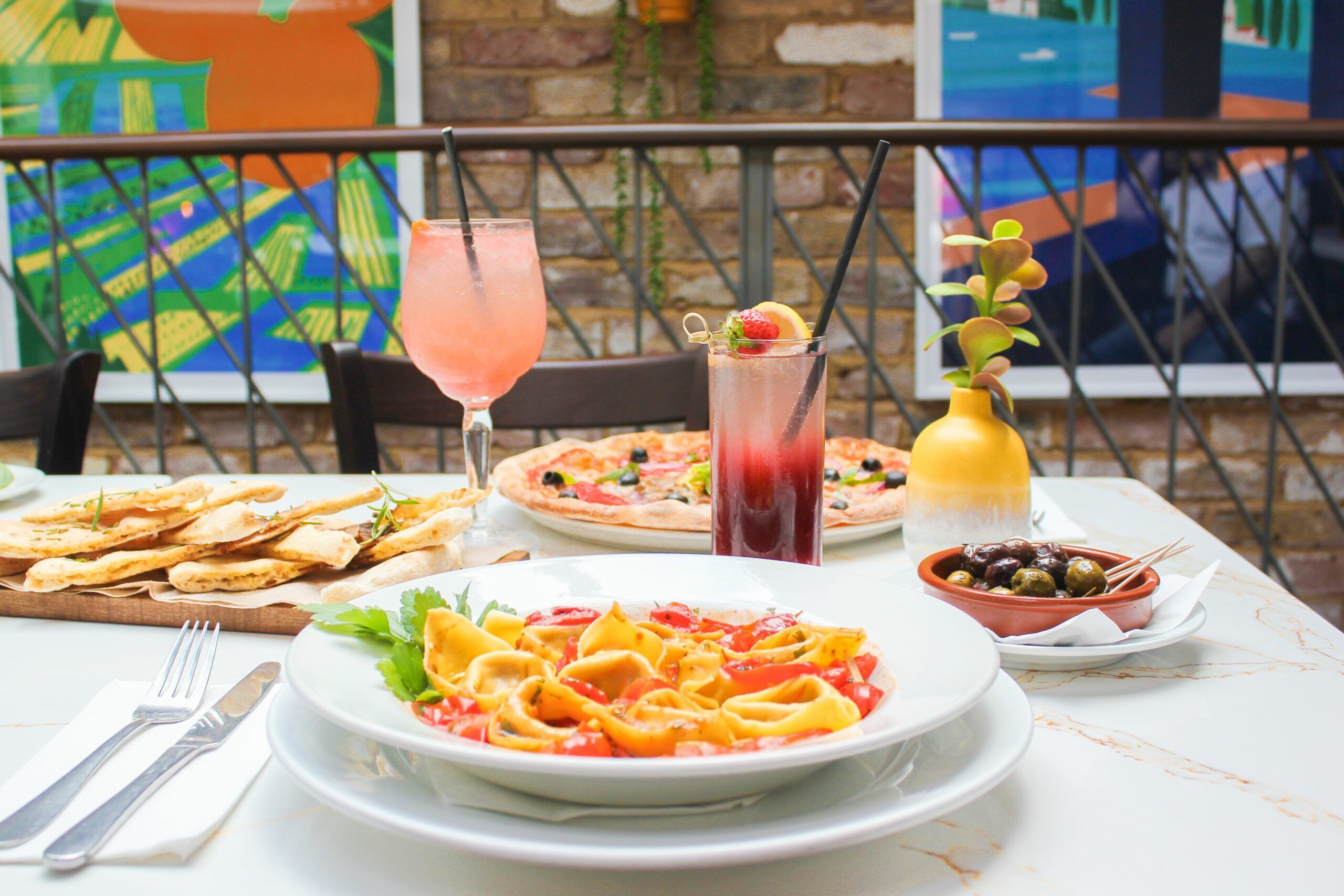 A marble table in Al Duomo Brighton is laid with a bowl of pasta, a pizza, some garlic bread, olives and two cocktails, one is pink and in a stem glass, the other is red and clear in a highball glass