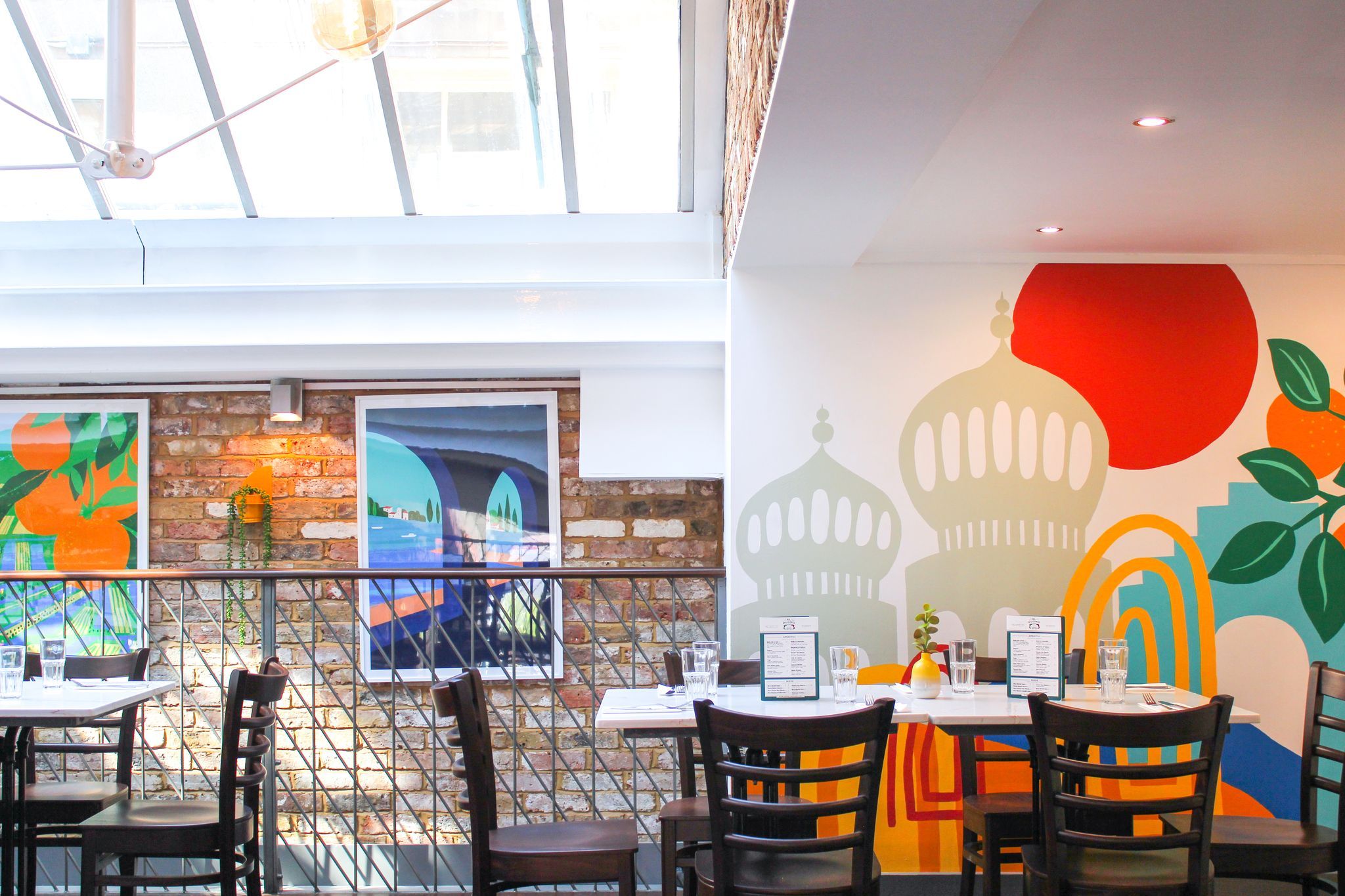 interior shot of Al Duomo Brighton. Wooden chairs arranged around square white marble tables. One wall is decorated with a mural of the Brighton Pavillion, another is exposed brick hung with pictures in frames. There is a large glass roof making the space very light.