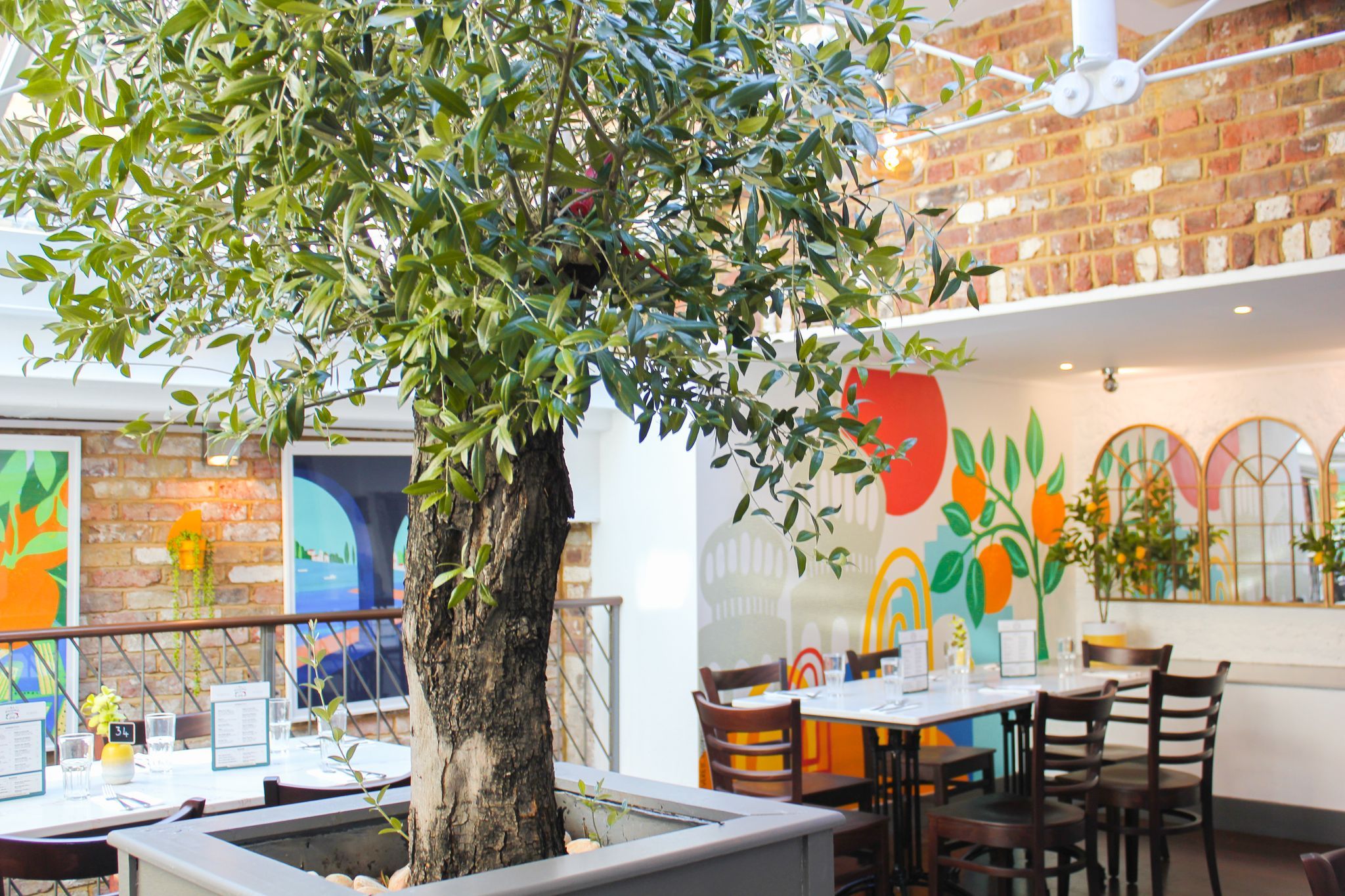 colorful interior at al duomo with a large tree growing in a planter underneath a glass roof. White walls are decorated with murals depicting Brighton pavilion and with arched mirrors. Exposed brick walls are hung with colourful prints in frames