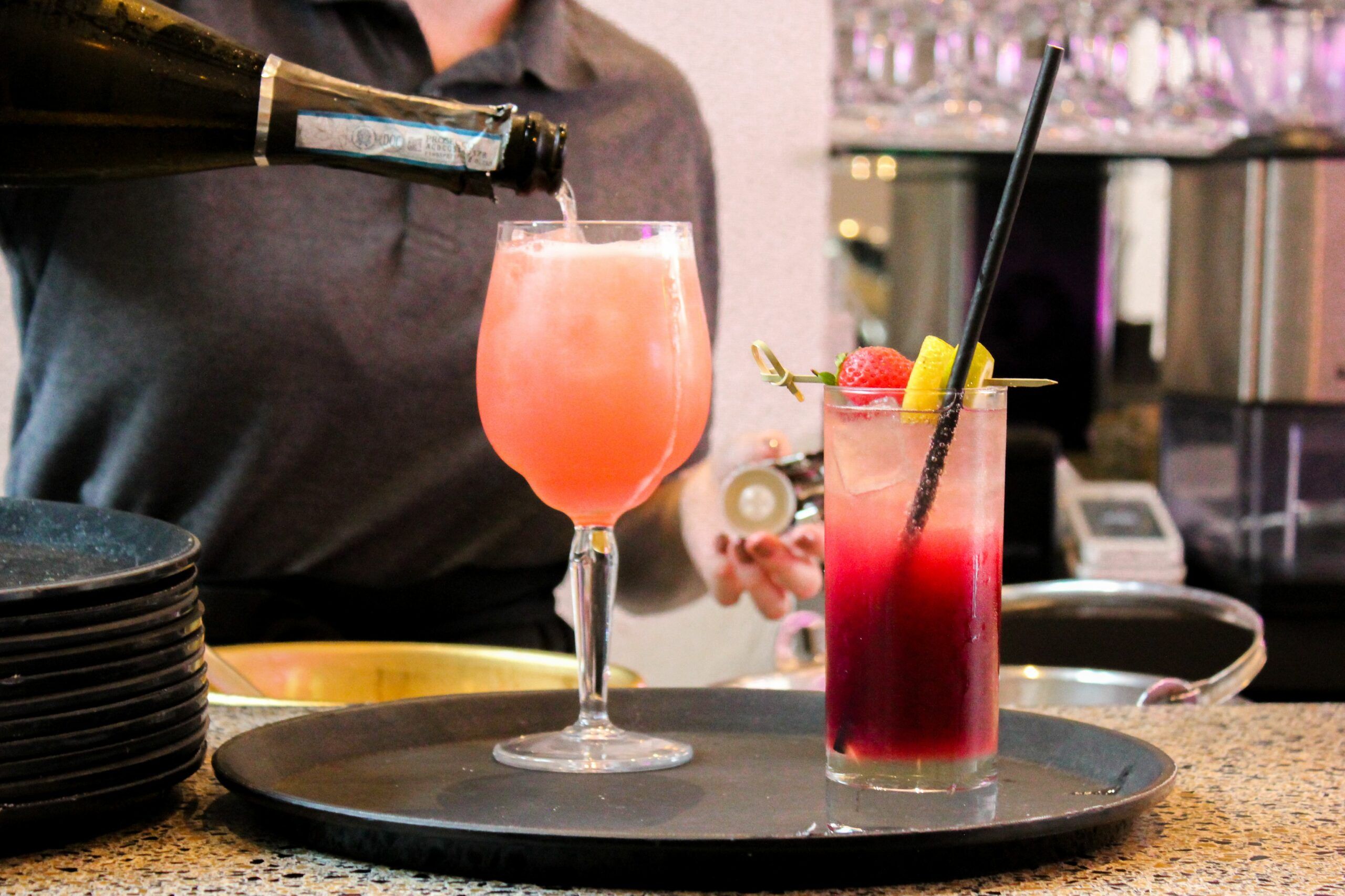 Two cocktails on a black tray, the left hand cocktail is being topped up by a server from a bottle of prosecco.