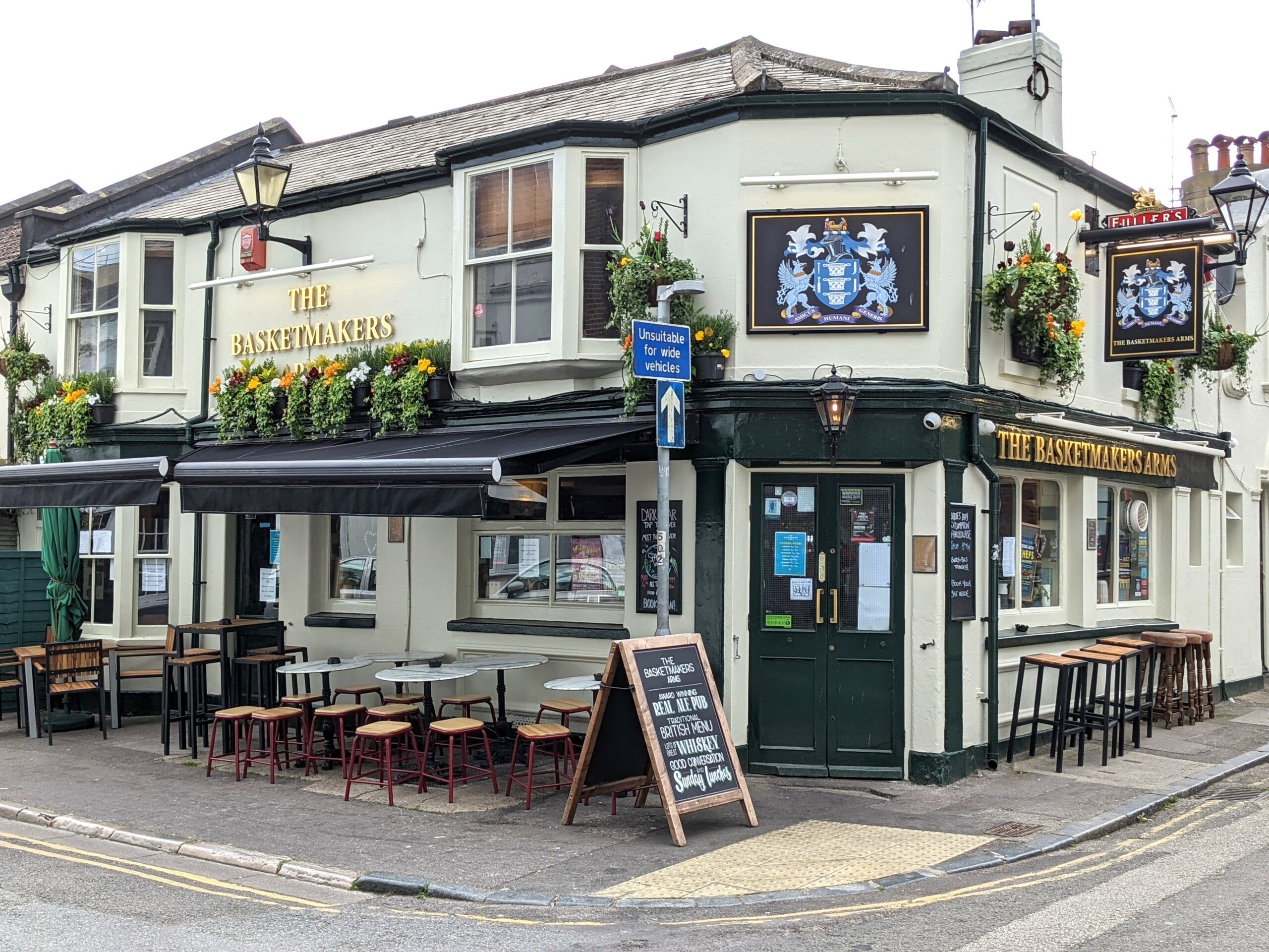 exterior of the basketmakers arms pub Brighton. Situated in the North Laine. It has a creamy white facade with black woodwor. the name of the pub is in gold. Tables and chairs are outside. 