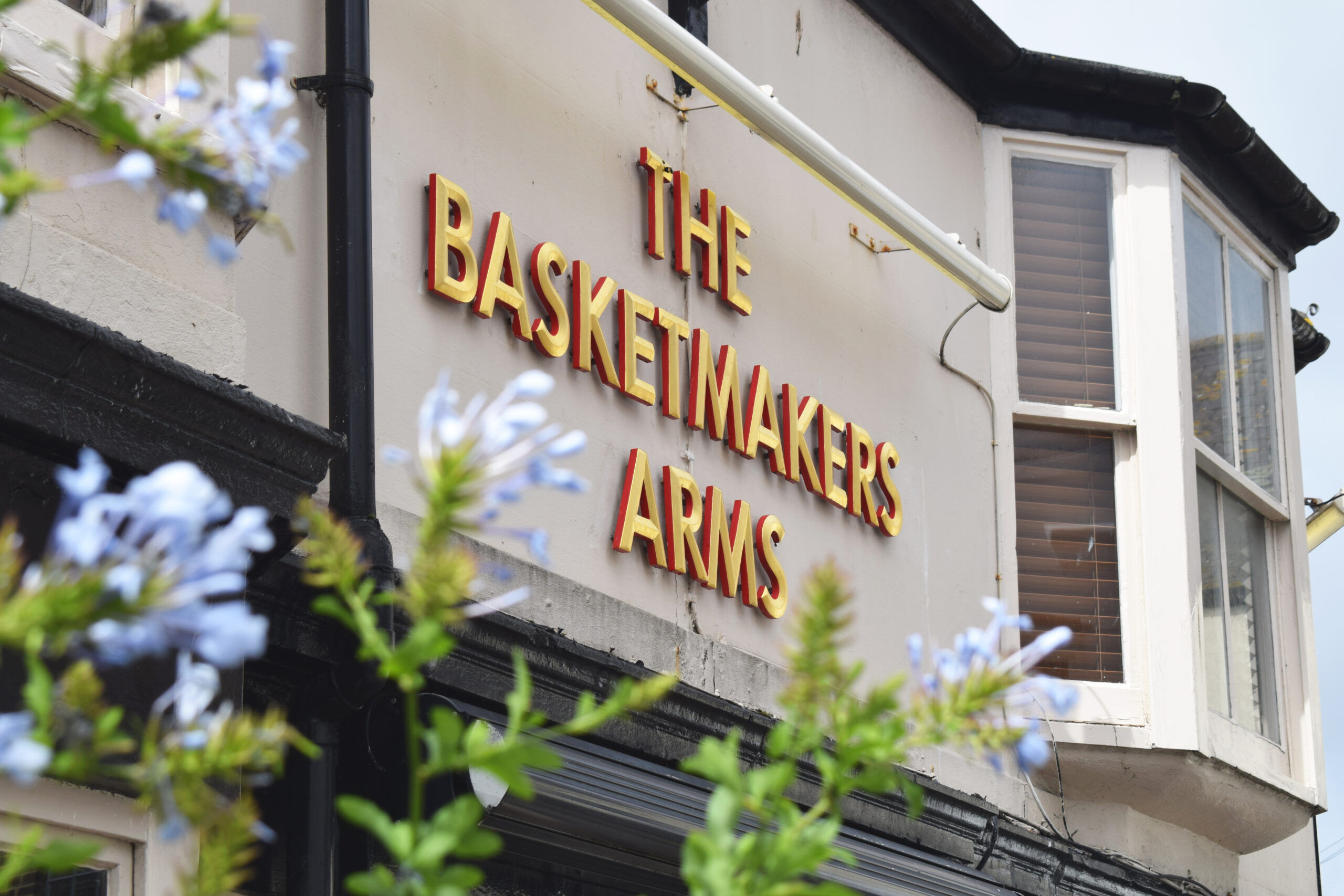 The Basketmakers Arms Brighton. Name of the pub written in gold with a creamy white facade. In the foreground you can see the pubs window box flowers. 