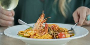 Seafood pasta served in a large white dish and a person holding a glass of white wine in the background.