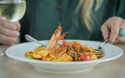 Seafood pasta served in a large white dish and a person holding a glass of white wine in the background.