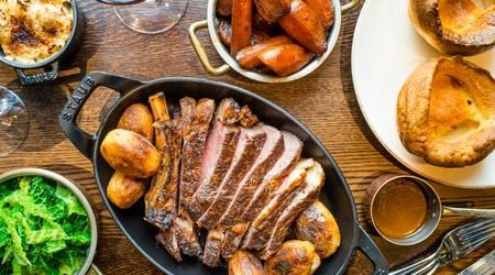 Overhead shot of a table full of roast dishes including a large bowl of sliced meat with potatoes, carrots, greens and Yorkshire puddings.