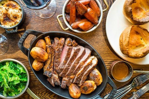 Overhead shot of a table full of roast dishes including a large bowl of sliced meat with potatoes, carrots, greens and Yorkshire puddings.